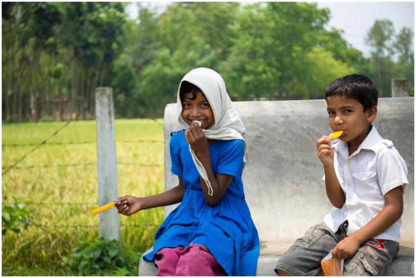 Two children enjoy ice pops while sitting on a ben