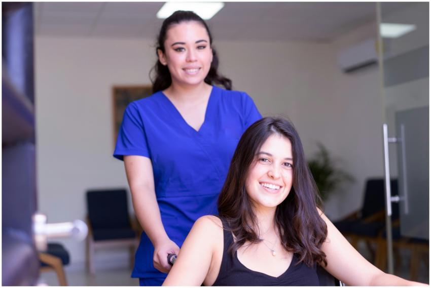 A nurse in blue scrubs assists a smiling patient i
