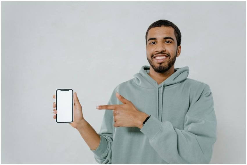 A smiling man in a hoodie points at a smartphone w