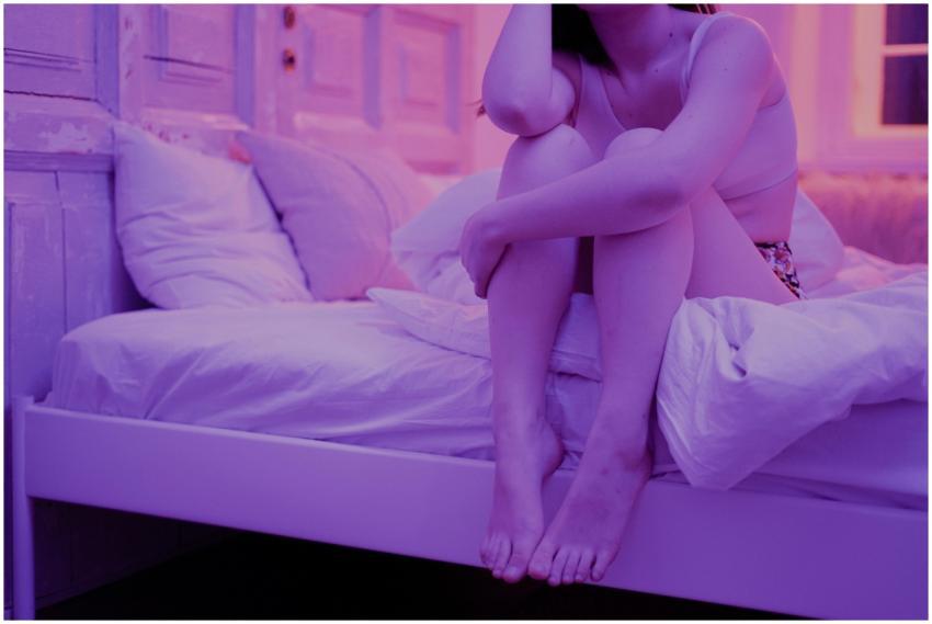 A woman sits on a bed in a dimly lit room, creatin