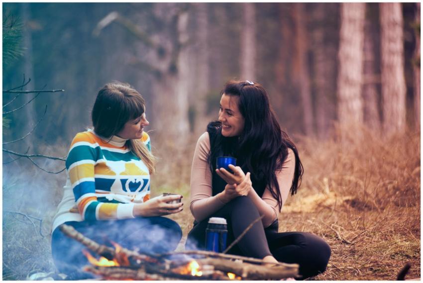 Two women enjoy coffee by a campfire in a forest,