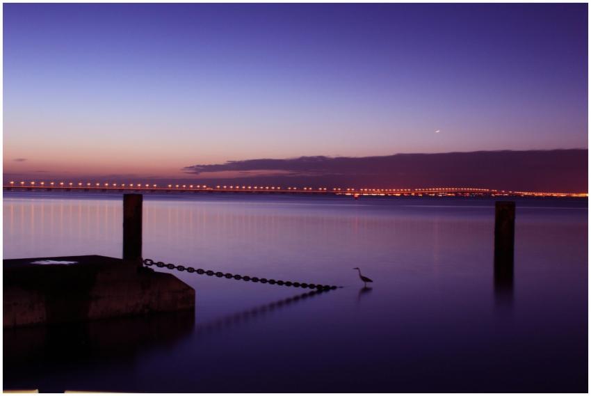 A tranquil seascape at dusk with a bridge and a he