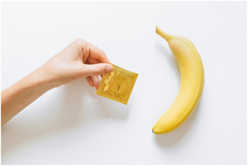 A hand holding a condom next to a banana on a whit