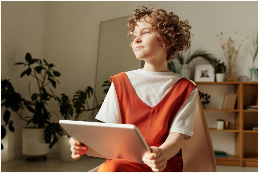A young boy sits indoors holding a tablet, deep in