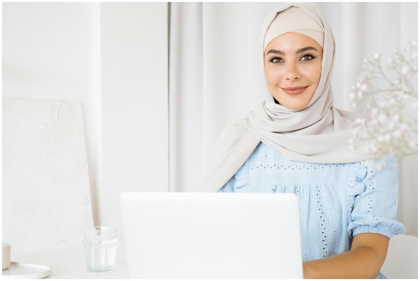 A smiling woman in a hijab using a laptop indoors