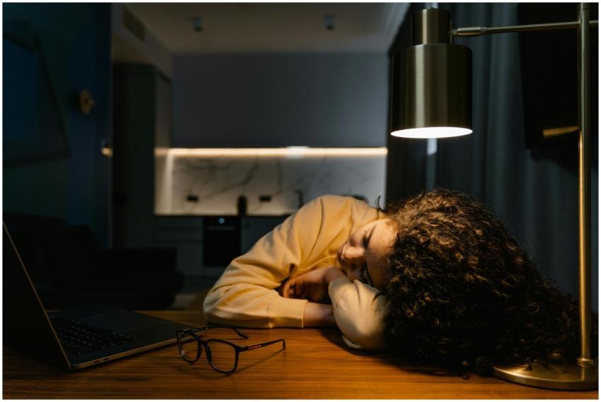 Exhausted woman sleeps at her desk under a lamp wi