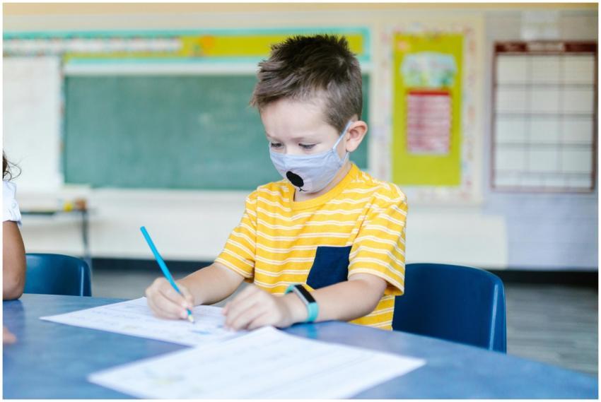 Child in a classroom drawing while wearing a face