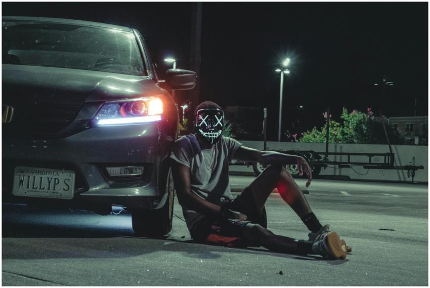 A person in a glowing mask sits beside a car at ni