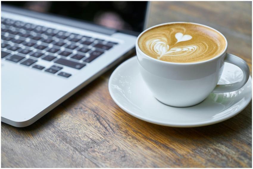 A cup of latte with intricate art beside a laptop