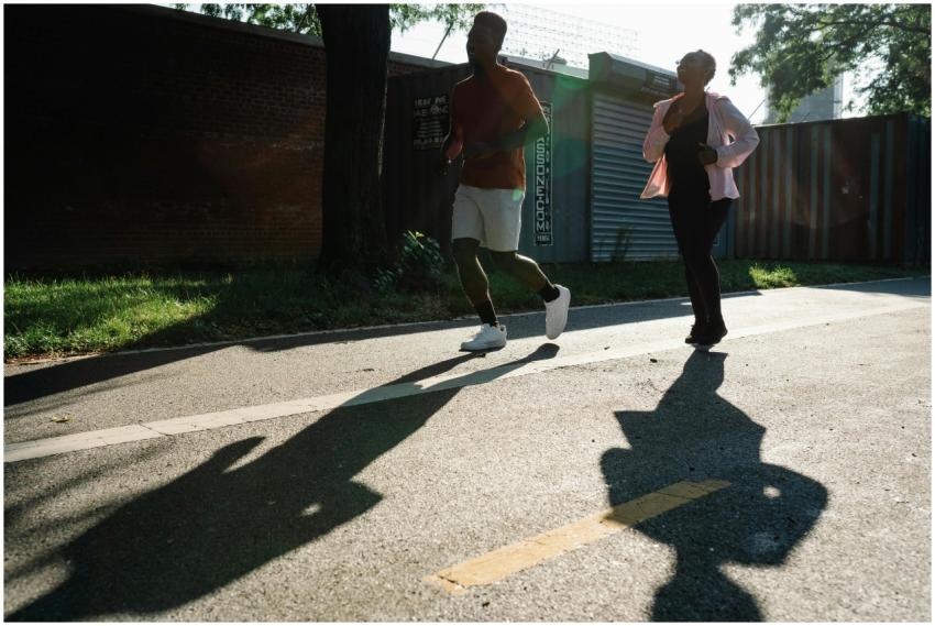 Two adults jogging on a sunlit sidewalk, casting d