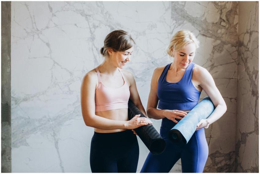 Two women in activewear holding yoga mats, smiling