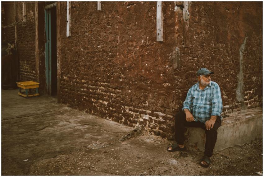 Elderly man sitting against a rustic wall in Ras E
