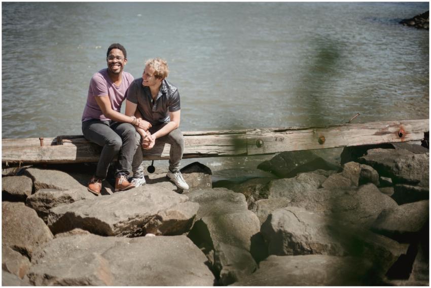 Two men laughing and cuddling on rocks by a rivers