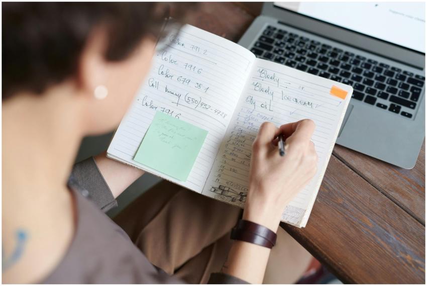 An adult writing in a notebook with a laptop on a