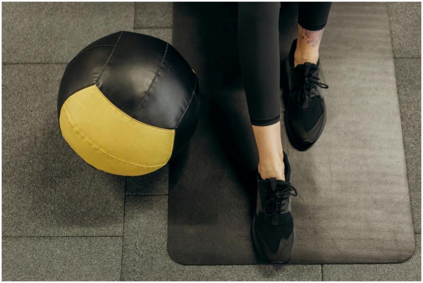 A woman sits on a gym mat with a medicine ball, re
