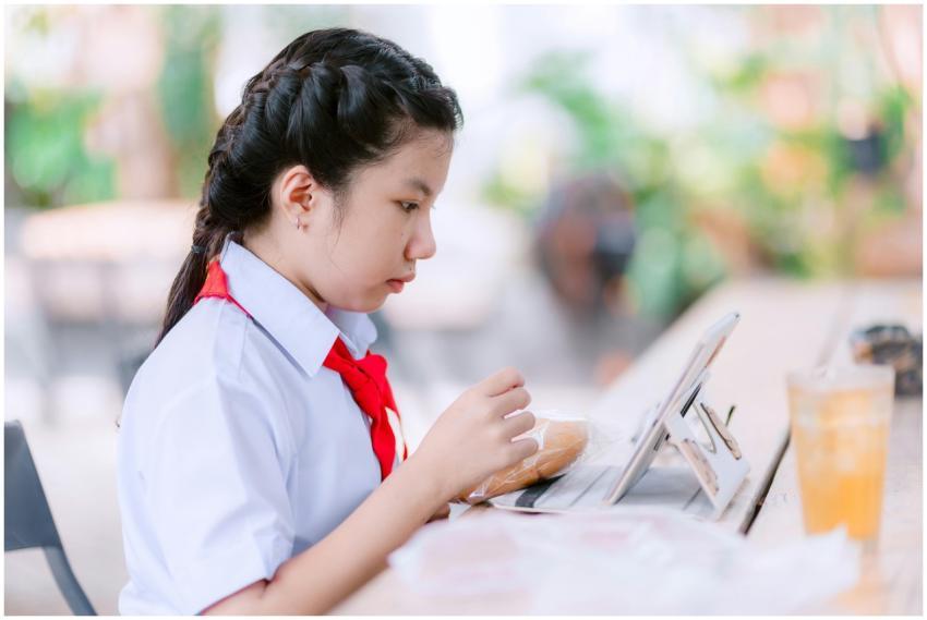 Schoolgirl studies on tablet with snack outdoors,