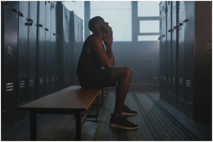 A man sits thoughtfully on a bench in a gym locker