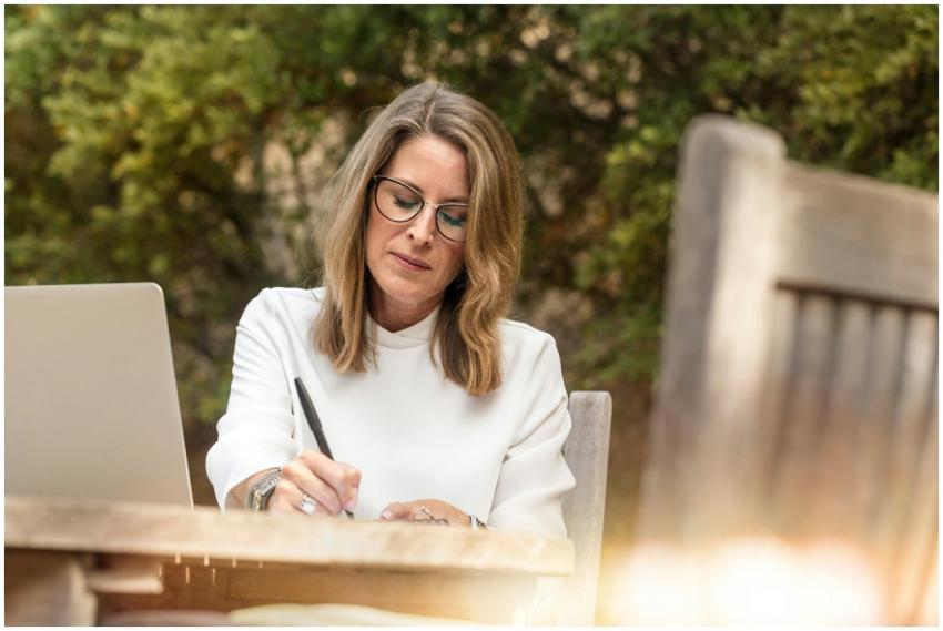 A woman in glasses writing and working on her lapt