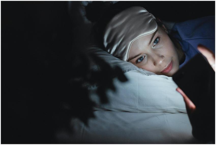 A woman in bed wearing a sleep mask uses her phone
