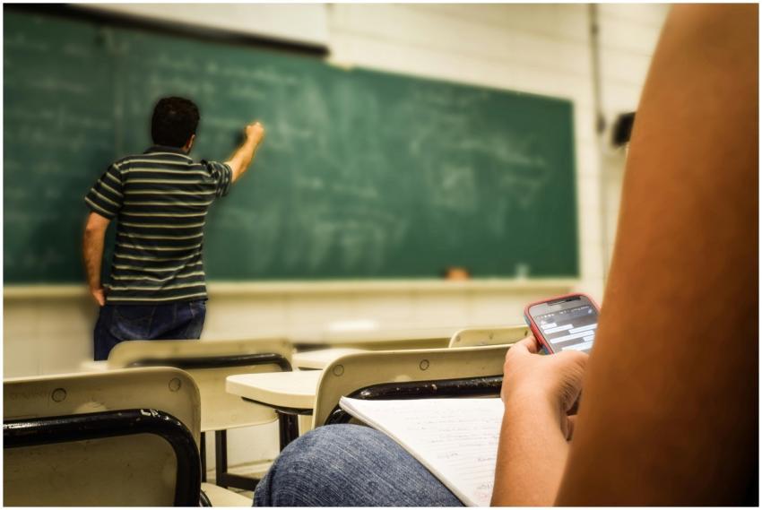 Student texting in a classroom while teacher is wr