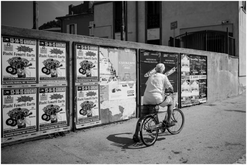 Elderly man biking past a wall of posters in a cit