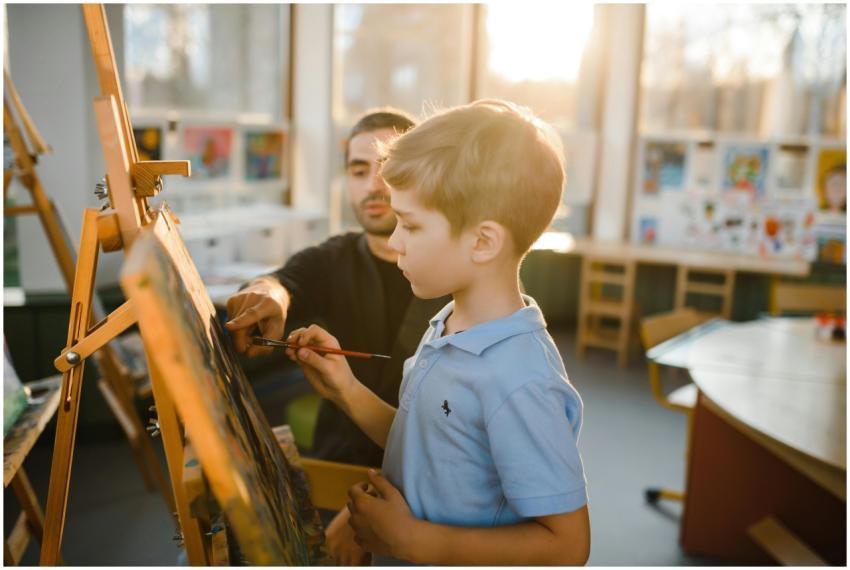 A young boy painting on an easel with a teacher as
