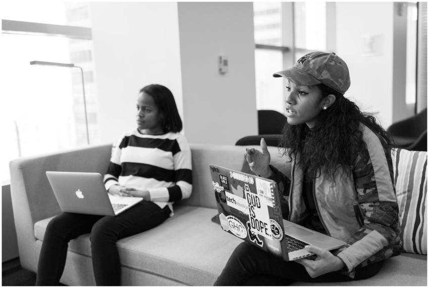 Two women seated indoors with laptops, engaged in