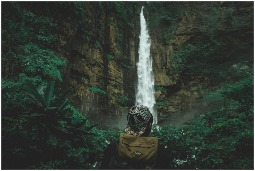 Person admiring a tall waterfall in a lush, green