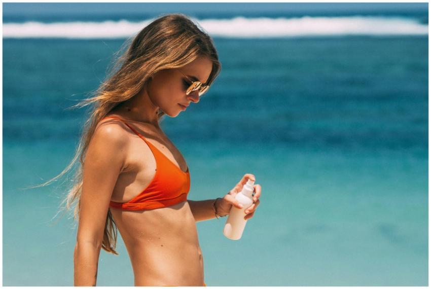 A young woman in a bikini applies sunscreen by the