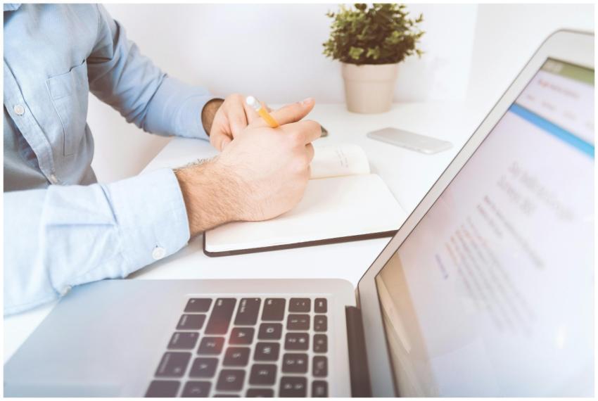A man writing in a notebook on a desk with a lapto