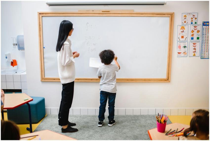 A teacher assists a young boy writing on a whitebo