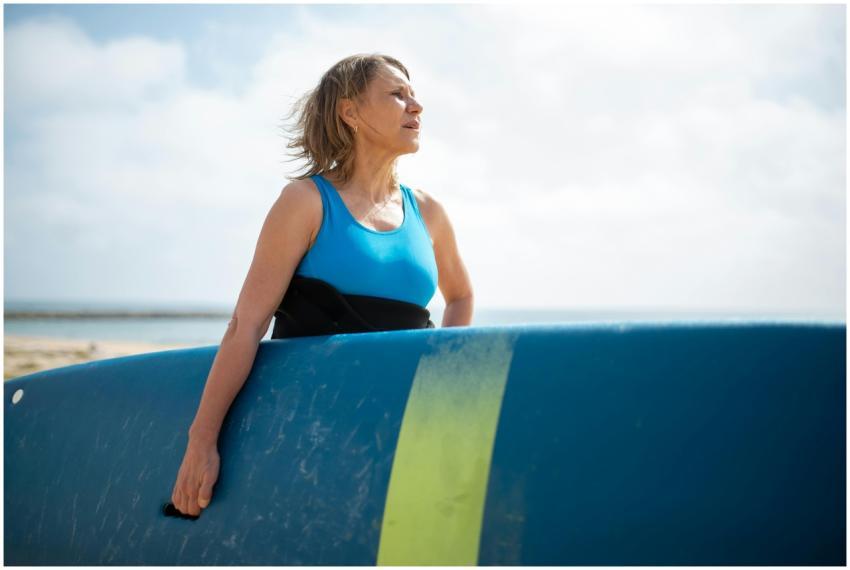 A senior woman in a wetsuit holding a surfboard, e