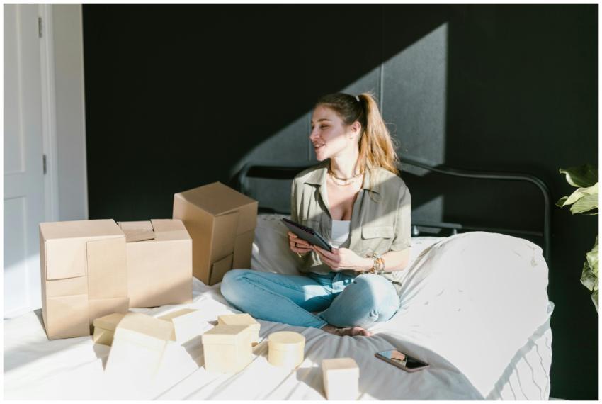 Woman sitting on bed with boxes and tablet, runnin