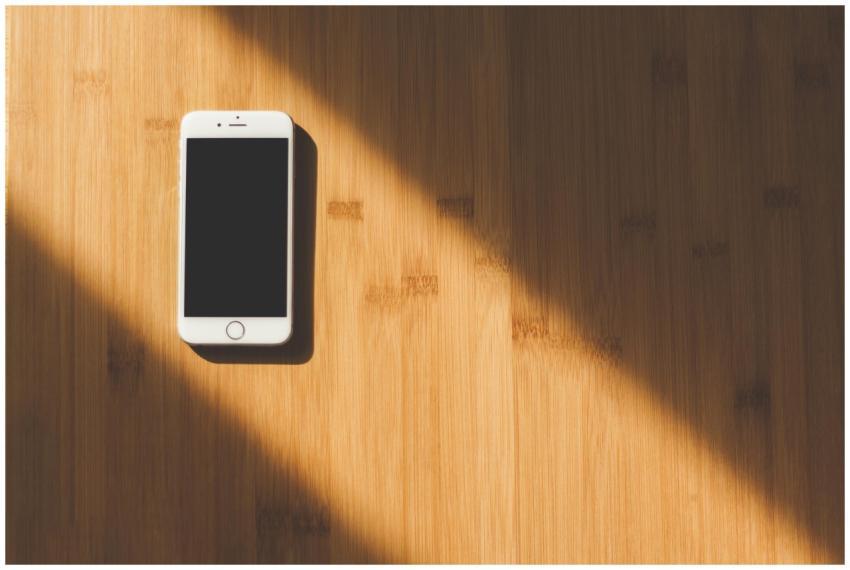 A sleek white smartphone lies on a sunlit wooden t