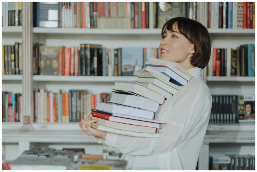 A woman joyfully holds a pile of books in a librar