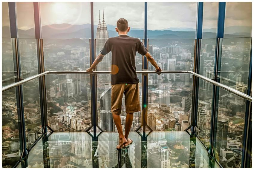 A man stands barefoot on a glass deck high above K