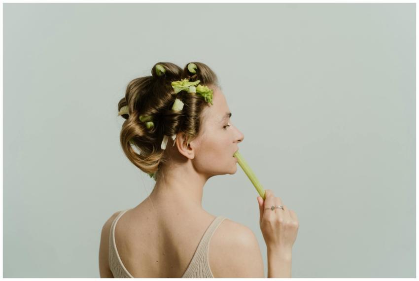 A woman with celery in hair rollers, biting a cele