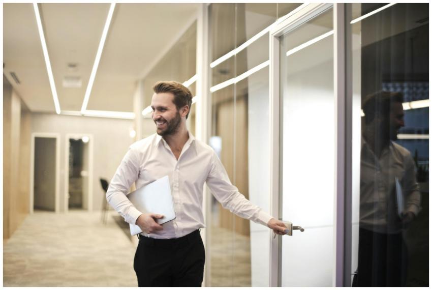 A smiling man holds a laptop while standing in a m