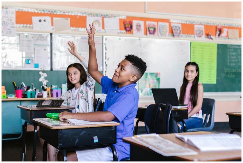 Children in a lively classroom raising hands durin