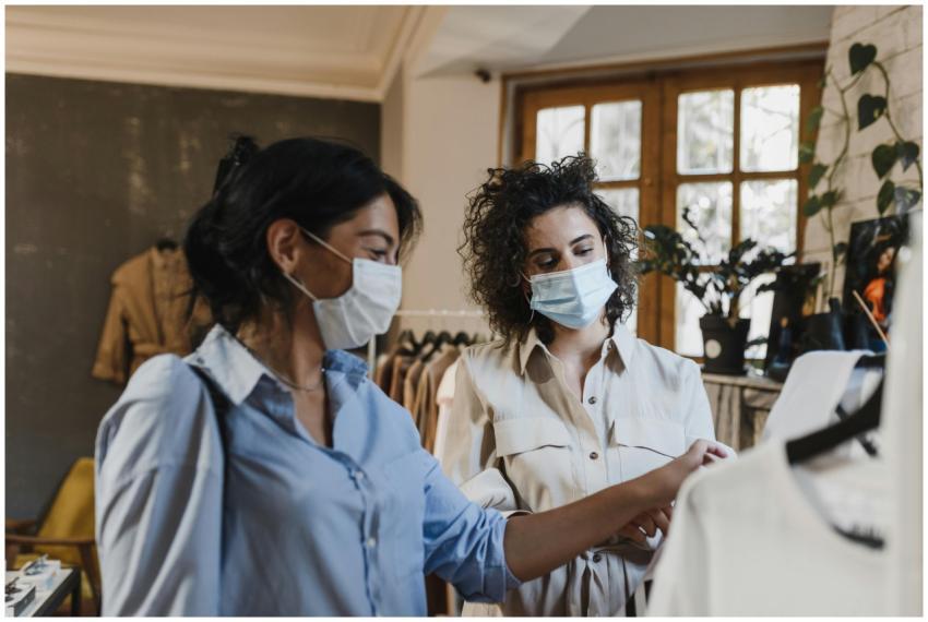 Two women browse clothing racks while wearing face