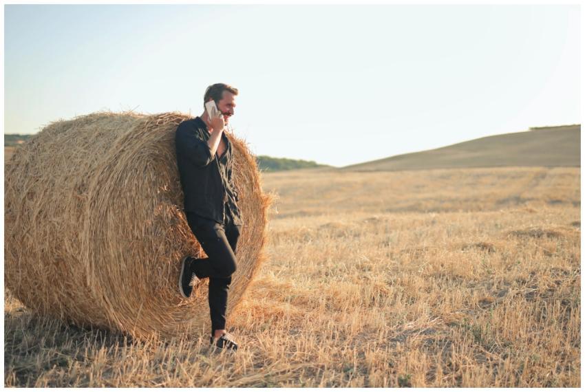 Adult man on phone in rural field, leaning on hay