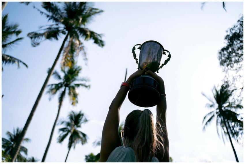 A woman lifts a trophy under palm trees, celebrati