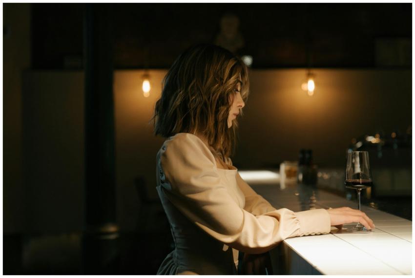 A stylish woman in a soft dress sits at a bar, sub