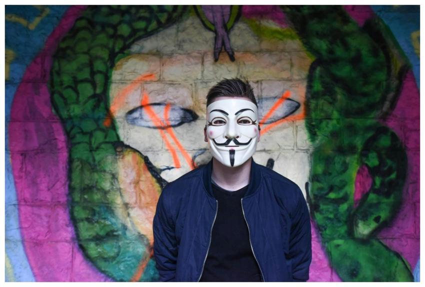 A person in a Guy Fawkes mask stands before colorf
