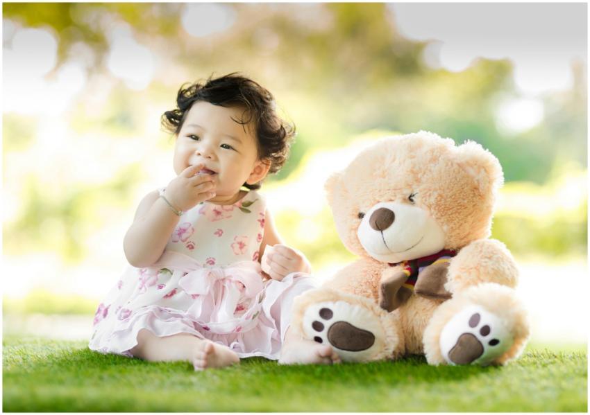 Smiling baby girl sits on grass with a plush teddy