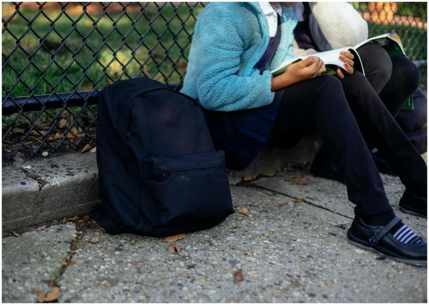 Children sit by a fence reading books, with backpa