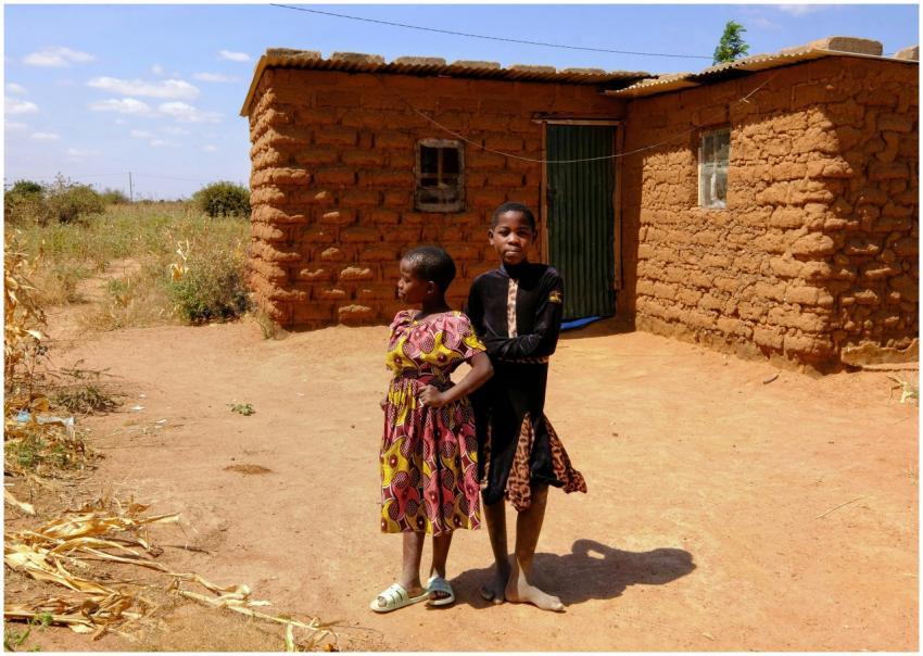 Two children stand in front of a mud house in rura