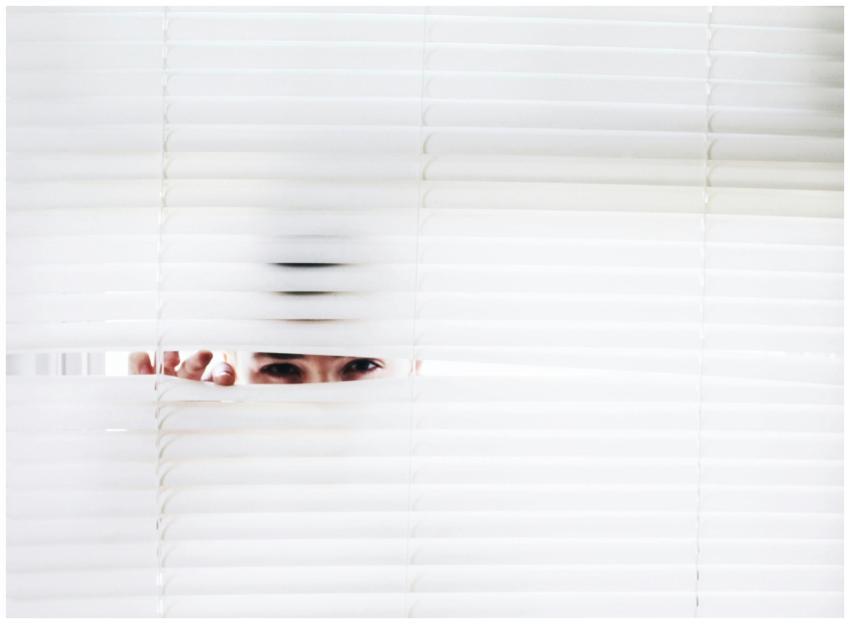 A person peeks through white blinds, creating an i