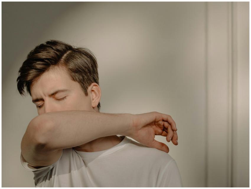 A young man sneezing into his elbow indoors, empha
