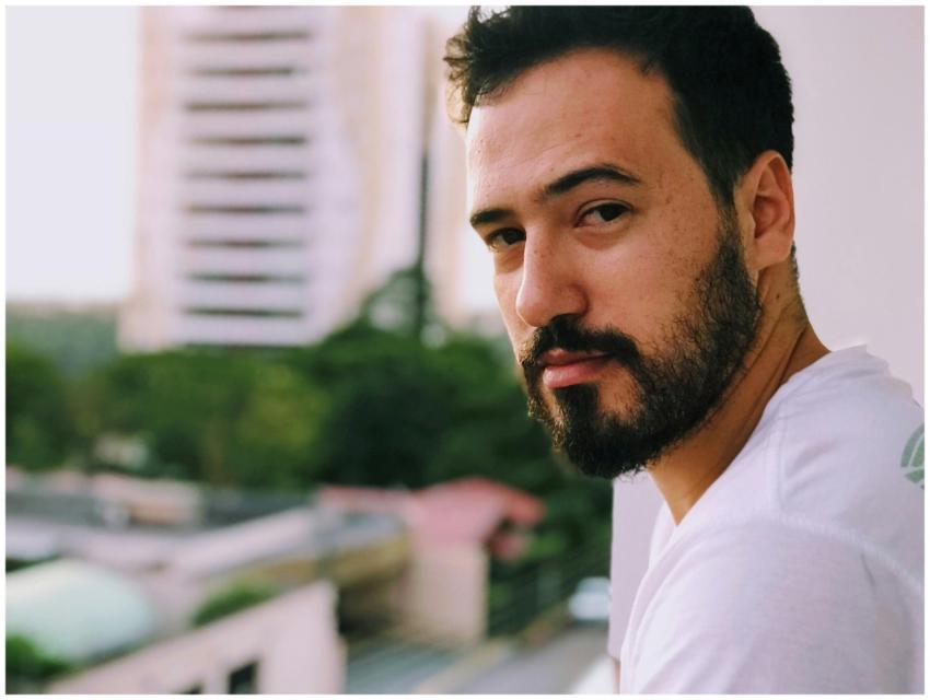 A serious-looking man with a beard stands on a bal
